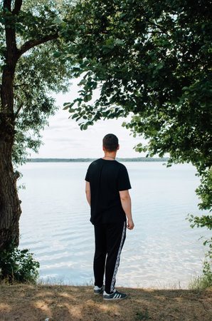 Back view of young man in casual clothes standing on shore near water and looking away, outdoors. Rear view of guy enjoying nature on sunny summer day.の写真素材