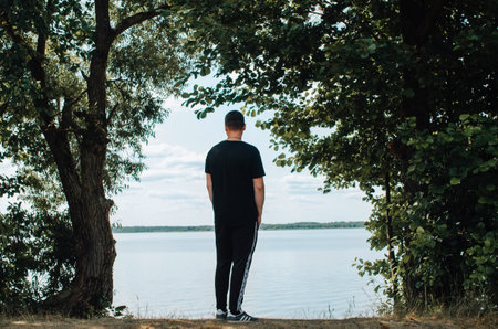 Young man in casual clothes standing on shore near water in tree frame and looking away at horizon...