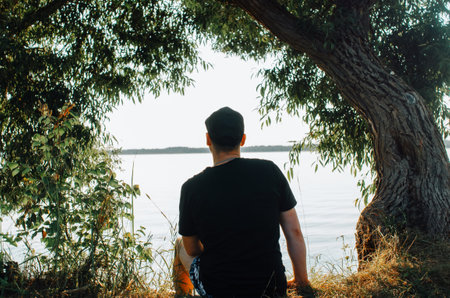 Caucasian young man in cap resting on shore near water in frame of trees and looking away at horizon outdoors, back view. Guy enjoying nature and sunset on sunny summer day, rear view.の写真素材
