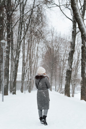 Back view of young woman in coat and knitted white hat enjoying walk in winter park, outdoors. Rear view of lonely girl walking along snowy alley. Vertical image.の写真素材