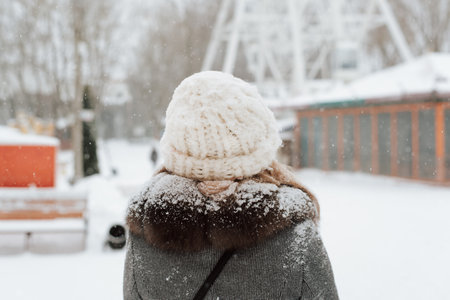 Back view of young woman enjoying snowfall, snowy weather in park in winter. Back view of girl in coat and cozy knitted white hat standing outdoors.の写真素材