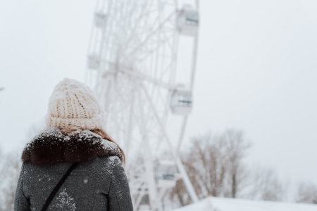 Back view of young woman in snow-covered coat and knitted white hat looking at ferris wheel attraction during winter walk in park, outdoors. Copy space.の写真素材