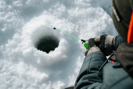 Close-up of fisherman sitting on frozen water near drilled hole in ice. Winter fishing, active...