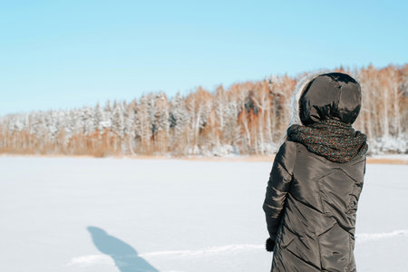 Back view of alone woman in hood and jacket standing on sunny frosty day in nature against backdrop of snowy forest and looking at winter landscape, copy space.の写真素材