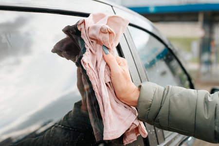 Car care concept. Female caucasian hand rubbing wet clean window of car door with rag after washing. Close-up, side view.の写真素材