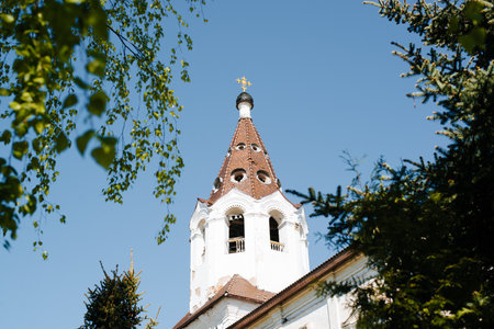 Close-up of white church with cross, an old monastery framed by trees on sunny day outdoors.