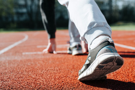 Close-up of woman practicing running in stadium, standing at low start on sunny day. Sportswoman exercising outdoors. Back view, selective focus on sneaker.の写真素材
