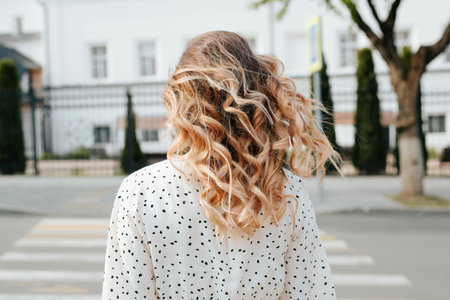 Close-up of stylish blond woman with curly hairstyle wearing polka dot dress walking on pedestrian crossing on street, back view.の写真素材