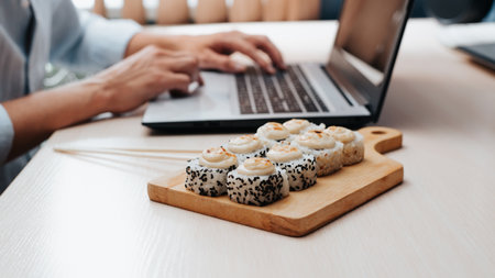 Close-up of man working on laptop in home office while sitting at table with meal of traditional Japanese cuisine, side view. Selective focus on baked hot sushi on wooden board.の写真素材