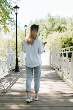 Rear view of stylish female student walking on bridge in park holding smartphone and laptop....