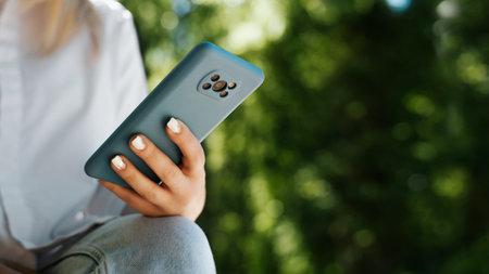 Close-up of woman's hand holding smartphone on sunny day outdoors, copy space. Woman using mobile phone in park in summer.の写真素材