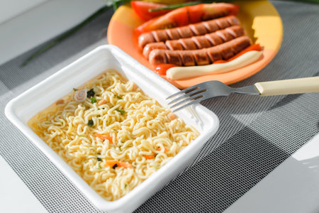 Close-up hot instant noodles, fried sausages with ketchup and mayonnaise, green onions and tomatoes on table.の写真素材
