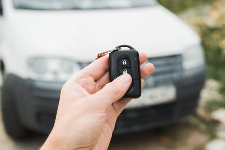 Close-up of human hand using car key remote control to unlock an alarm. Selective focus on remote control, white car in bokeh.の写真素材