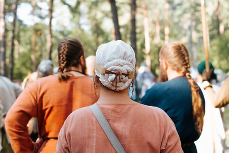 Back view of an unrecognizable woman with an ancient costume and a traditional scarf on her head standing in a crowd of medieval men outdoors.の写真素材
