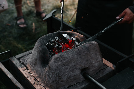 Blacksmith holding tongs and iron working in a medieval forge, close-up. Selective focus on hot coals.の写真素材
