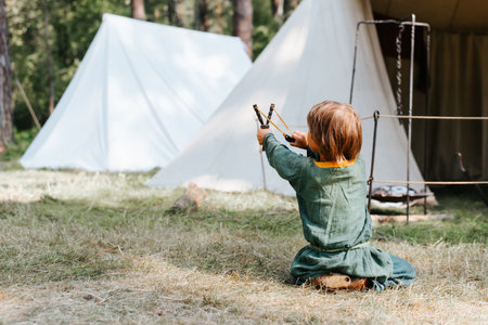 Little boy in medieval rustic clothes playing in forest. Child shooting from slingshot sitting on grass outdoors, back view.の写真素材