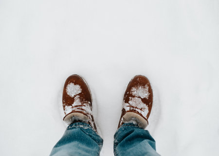 Man wearing winter leather brown boots and blue jeans standing in snow outdoors. Top view, close-up, copy space.の写真素材
