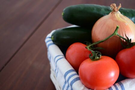 A bunch of a tomatoes in a basket with blurred cucumbers and a onion in the backgroundの写真素材