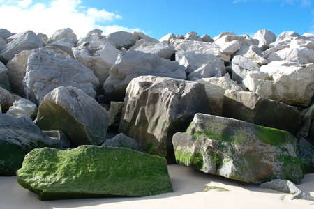 Sea breaker made with natural rock with algae growing on some of them and a blue sky and some puffy cloudsの写真素材
