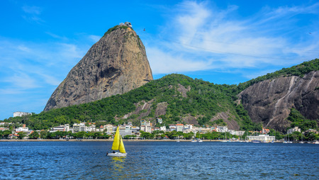 Yellow small sailing yacht and classic daytime scenic profile view of Sugarloaf Mountain, Pao de Acucar, standing above Botafogo Bay in Rio de Janeiro, Brazilの写真素材