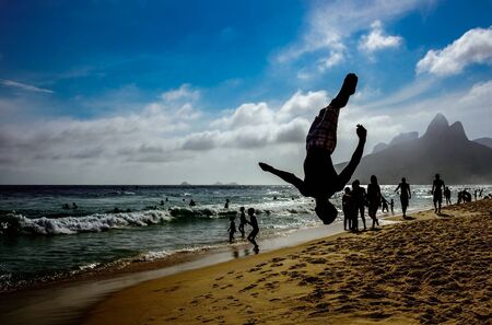 Silhouette of the man perfoming back somersault at Ipanema beach with silhouettes of swimming and walking people, blue sky, white clouds and Dois Irmaos Mountain at background, Rio de Janeiro, Brazilの写真素材