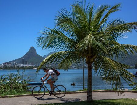 RIO DE JANEIRO, BRAZIL - OCTOBER 31, 2016: Cyclist riding by famous bicycle and running track at residential neighborhood Lagoa, Rodrigo de Freitas Lagoon and Dois Irmaos Mountain on the backgroundのeditorial素材