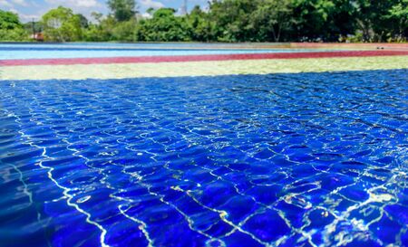 Light blue, red, yellow and blue striped mosaic floor of fountain covered with clear water at sunny day on the background of blurry green treesの写真素材