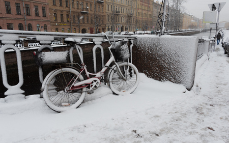 Red bicycle with baskets covered with snow leaning against a fence next to Griboyedov Canal at snowing winter dayの写真素材