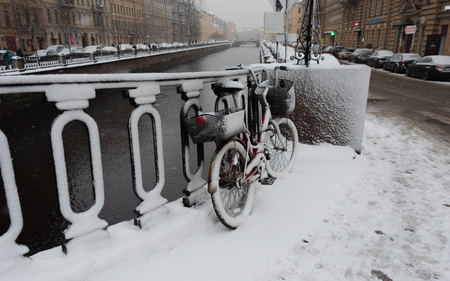 Red bicycle with baskets covered with snow leaning against a fence next to Griboyedov Canal at snowing winter dayの写真素材