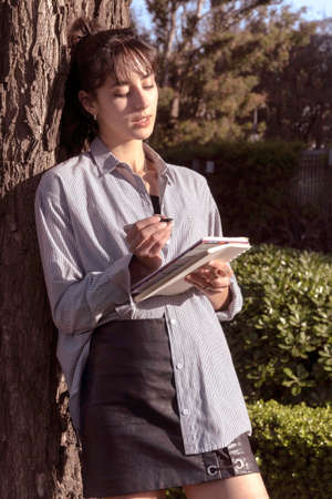Young woman writes in a notebook leaning against the trunk of a tree, and the afternoon sun...