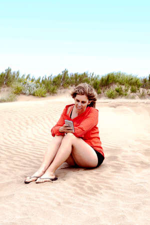 woman looks at her smartphone sitting on the sand at the seashoreの写真素材