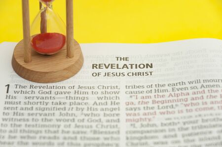 Red sand hourglass on top of open Holy Bible in the Book of Revelation of Jesus Christ chapter 1. Isolated on yellow background. Close-up. Horizontal shot.の写真素材