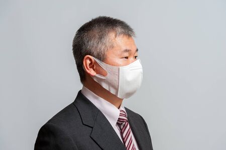 Side view of middle aged Asian man with suit and tie wearing white surgical mask for protection against coronavirus (COVID-19) and other infectious diseases. Isolated on white background. Looking to the side.の写真素材