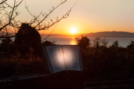 Holy Bible opened with directional light. Background with sea, golden sky and sunrise behind the mountains. Copy space. Horizontal shot.の写真素材
