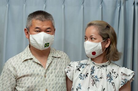 COVID-19 Pandemic - Front view portrait of middle age wearing couple DIY homemade surgical face mask with Brazil and Japan flag. Corona protection. Indoors.の写真素材