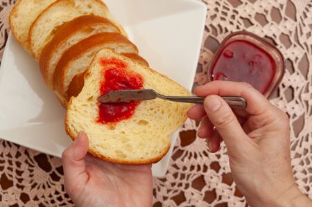 Traditional Brazilian homemade bread - Hands putting strawberry jam on bread. Crochet towel background. Close-up. Top view.の写真素材
