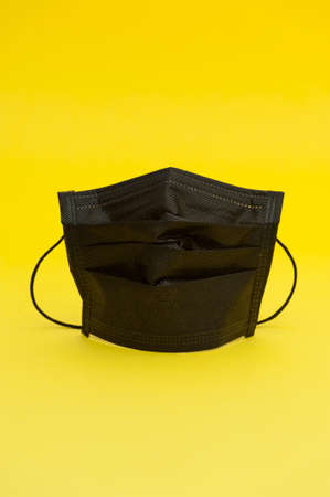 Black surgical mask for protection against Coronavirus COVID-19 and other contagious diseases. Isolated on yellow background. Vertical shot. Copy space.の写真素材