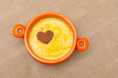 Homemade sweet corn pudding known in Brazil as Curau or Canjica Nordestina in ceramic bowl. Powdered cinnamon in the heart shape. Isolated on jute background. Copy space. Top view. Horizontal shot.の写真素材