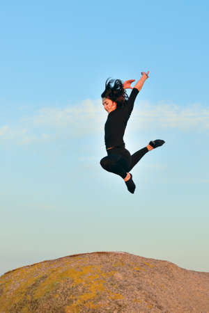 image of young woman doing gymnastic jump on a rock against a blue skyの写真素材