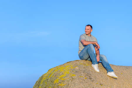 young athletic attractive man sitting on a rock against a blue sky background and smiling looking to...