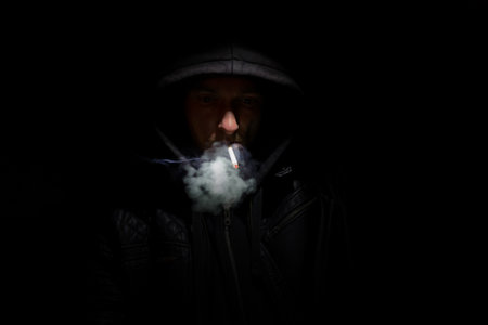A man is smoking a cigarette. He is wearing a black hoodie. The cigarette is lit and he is holding it in his mouthの写真素材