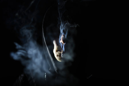 A man is smoking a cigarette. He is wearing a black hoodie. The cigarette is lit and he is holding it in his mouthの写真素材