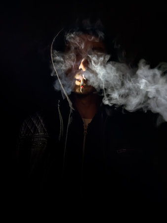 a man is smoking a cigarette. He is wearing a black hoodie. The cigarette is litの写真素材