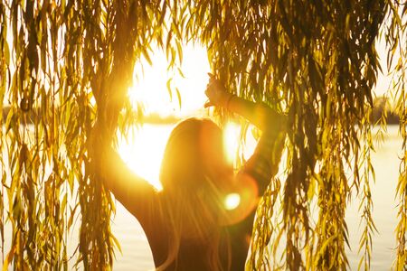 Autumn portrait of young girl with willow branches on the background of a lake at sunsetの写真素材