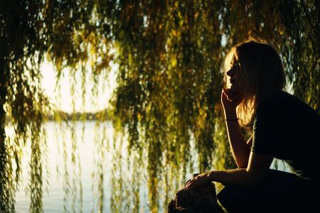 Autumn portrait of smiling young girl near willow on the background of a lake at sunsetの写真素材