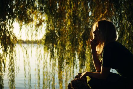 Autumn portrait of smiling young girl near willow on the background of a lake at sunsetの写真素材