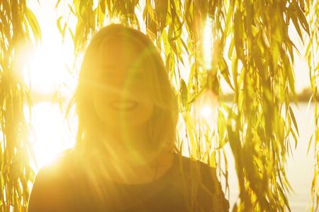 Autumn portrait of young happy smiling girl with flying hair with willow branches on the background of a lake at sunsetの写真素材