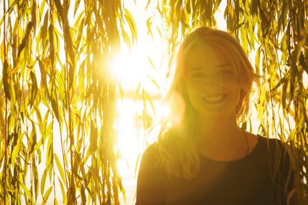 Autumn portrait of young happy smiling girl with flying hair with willow branches on the background of a lake at sunsetの写真素材