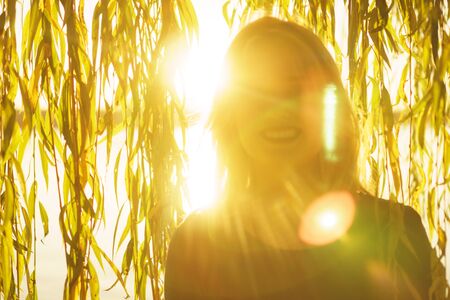 Autumn portrait of young happy smiling girl with flying hair with willow branches on the background of a lake at sunsetの写真素材