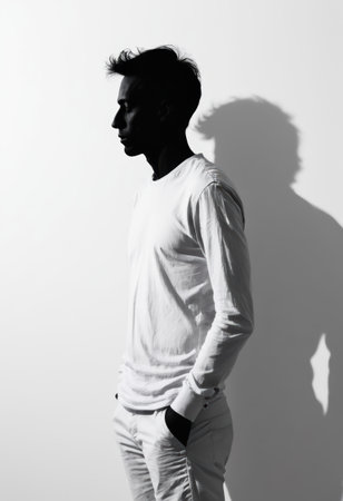 A man in a white long-sleeve shirt stands silently in profile, deep in thought, against a minimalist white backdrop, creating a striking contrast with his silhouette at dusk.の素材